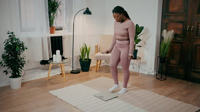 Young hopeful african american woman checking weight on floor scales and enjoying weight loss, dancing at home