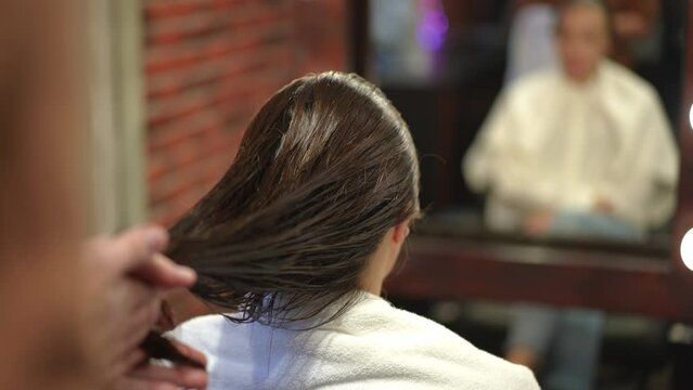 Close-up unrecognizable hairstylist applying hair lotion on long brown female hair. Young Caucasian man doing hairstyling in beauty salon indoors. Haircare and beauty concept