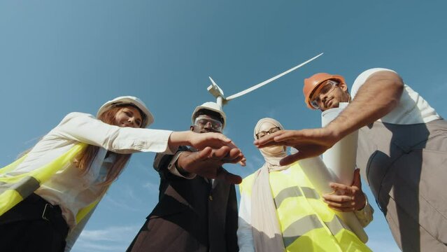 Men and women in safety helmets having successful meeting outdoors. Group of four multicultural partners stacking hands together and smiling on camera while standing among wind turbines