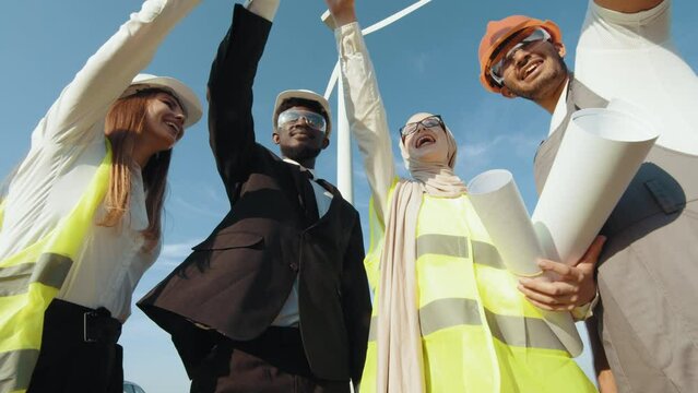 Group of four multicultural partners stacking hands together and smiling on camera while standing among wind turbines. Men and women in safety helmets having successful meeting outdoors