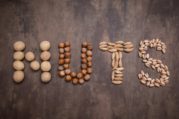 Word nuts from different type of nut. Walnut, hazelnut, almond, pistachio on brown abstract background, top view, flatlay	
