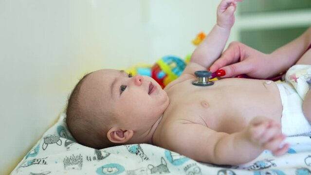 Baby doctor checks little boy’s health with stethoscope. Kid looks up cheerfully, waving hands, legs and smiling to the pediatrician. Side view.