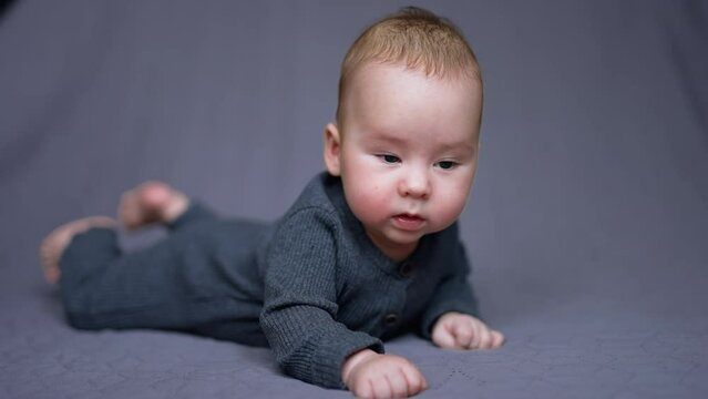 Sweet little boy in grey clothes lies on his belly. Healthy child looks around and then up. Grey blurred backdrop.