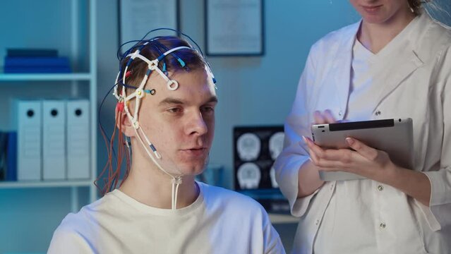 EEG topographer for examining brain, doctor making electrogram of scalp activity. Researching of brain work man wearing electroencephalography headset, nurse writing notes in gadget tablet.