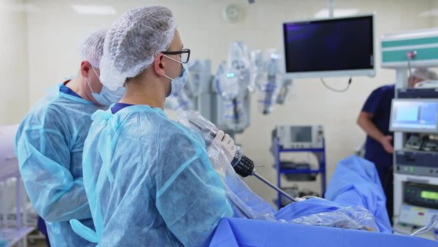 Younger male surgeon holds long device attached to a patient. Chief doctor stands beside. Highly-equipped operational theatre at backdrop in blur.
