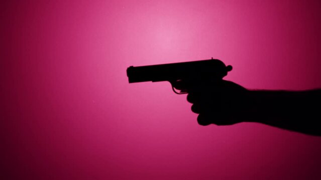 Man holding gun close-up, shadow silhouette of hand with pistol on pink neon background. Horror film and killer attack. crime concept, using weapon.