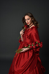 Fototapeta premium portrait of pretty female model with red hair wearing glamorous historical victorian red ballgown. Posing with a moody dark background.