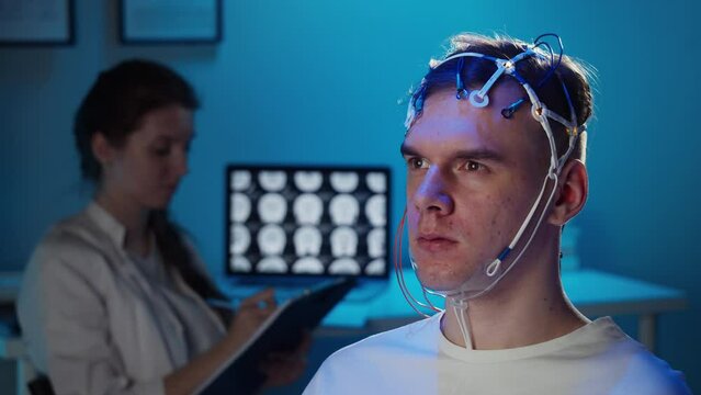 EEG topographer for examining brain, doctor making electrogram of scalp activity. Researching of brain work man wearing electroencephalography headset and watching video, nurse writing notes.