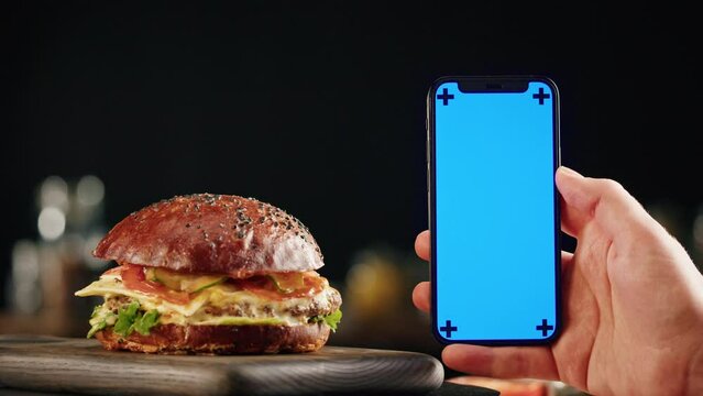 Tasty burger and smartphone with blue chroma key screen close-up, cooked juicy sandwich, fast food delivery application, recipe of preparing homemade hamburger with meat and fresh vegetables.