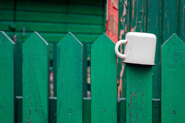 A white mug hangs on a green fence.