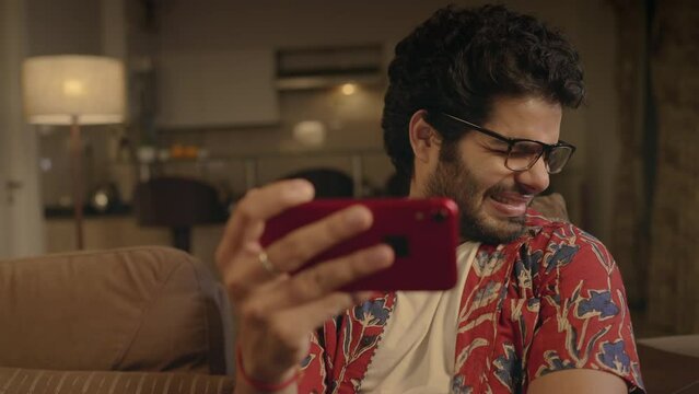 young Indian Asian man with specs sitting on a couch and making facial expressions or closing eyes while watching a violent movie or disturbing serial content on Ott platform over a Smartphone at home