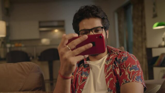 close up shot of a young happy Indian Asian attractive male guy wearing specs sitting on a couch and smiling while watching a Comedy Movie or video on Ott platform using a mobile phone at home night