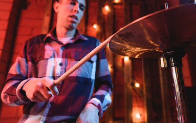 Crop talented male musician in checkered shirt playing drums with drumsticks while performing song on stage with glowing lamps during concert