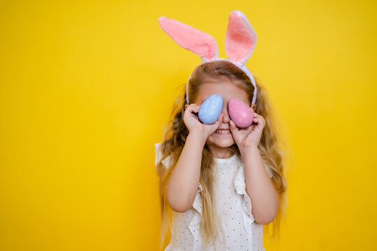 beautiful smiling blonde girl with bunny ears holding an easter egg in her hands, closes eyes, on a yellow background, kid celebrate easter.