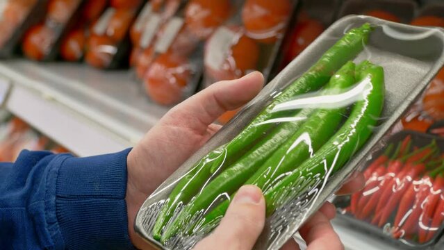 A man in a grocery store chooses hot jalapeno peppers. A customer in the market at the vegetable counter holds a package of fresh green hot peppers in his hands. Buying vegetables for dinner.