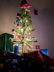 Green Christmas tree with lights decorated with typical figures of the date, with red balls, with mini gifts, and a red bow at the top of the tree with a white background. Vertical photography.