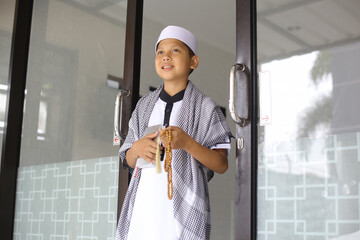 Smiling muslim boy holding al quran and tasbih and walking out from mosque