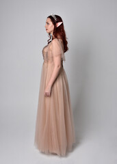 Fototapeta premium Full length portrait of pretty female model with red hair wearing glamorous fantasy tulle gown and crown. Posing with gestural arms on a studio background