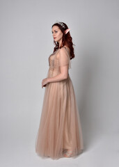 Full length portrait of pretty female model with red hair wearing glamorous fantasy tulle gown and crown.  Posing with gestural arms on a studio background