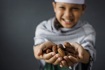 Selective focus of smiling muslim kid giving handful of dates on gray and blurred background
