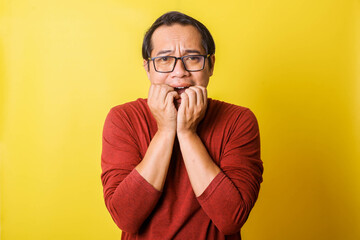 Casual man in glasses with scared expression biting fingertip isolated on yellow background