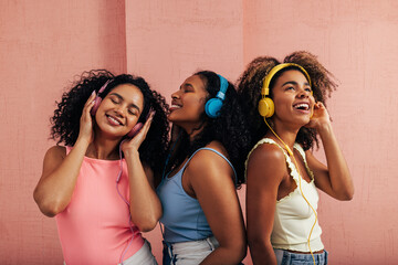 Three beautiful women standing together and listening to music. Girlfriends with headphones wearing...