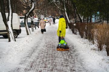 Mom carries a child along a snow-covered city road on a sled on a winter evening