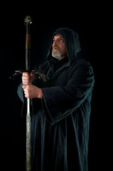 Portrait of courageous monk-warrior in black cloak with sword in hands on black background. Historical medieval templier fantasy