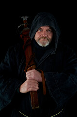 Portrait of courageous monk-warrior in black cloak with sword in hands on black background....