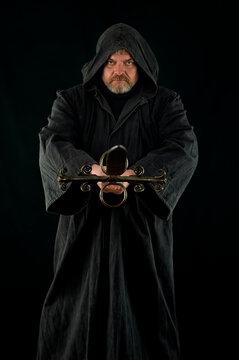Portrait of courageous monk-warrior in black cloak with sword in hands on black background. Historical medieval templier fantasy