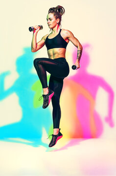 Sporty girl jumping with dumbbells. Photo of  muscular woman in black sportswear on white background with effect of rgb colors shadows. Dynamic movement. Sports motivation and healthy lifestyle