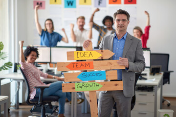 A male manager is holding a sign with company slogans and posing for a photo in the office with his colleagues. Employees, job, office