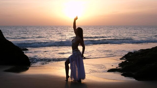 Sexy woman silhouette at beach at sunset. Lady touching sun with hand. A beautiful woman performing a sensual belly dance in the sand on the beach. Spectacular landscape with blanks. Silhouette.