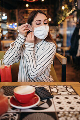 Beautiful young adult woman sits down in a nice restaurant and takes off her protective face mask. She is alone and serious. Coronavirus pandemic and social distancing problems.