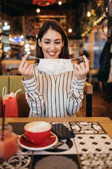 Beautiful young adult woman sits down in a nice restaurant and takes off her protective face mask. She is alone and serious. Coronavirus pandemic and social distancing problems.