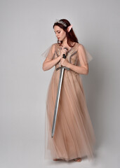  Full length portrait of pretty female model with red hair wearing glamorous fantasy tulle gown, crown. Holding a sword weapon, on studio background. © faestock