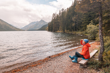 A traveler girl is sitting on a bench and enjoying a great panoramic view of a mountain lake in Altai. Hiking in the national park and solitude in nature