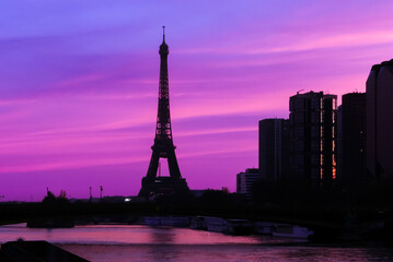 View on the Eiffel tower with group of modern buildings in front of the water of Seine river. Dramatic sky with colorful clouds. Silhouette of a cityscape at sunrise.