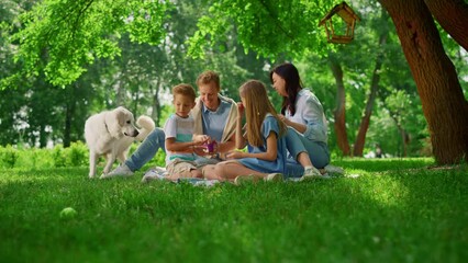Happy family feed white dog on weekend picnic. People rest with pet in park.
