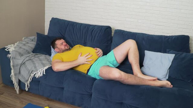 Plump man with beard in yellow T-shirt is lying on the bed at home and stroking big sick stomach. Bloating, overeating, cramps and pain, diarrhea. Proper nutrition