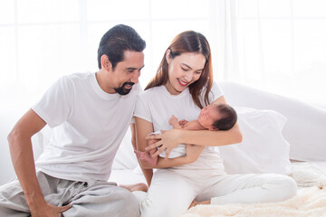 Selective focus of Asian man and woman holding newborn baby in bed at home. Happy family smiling with adorable infant, young parents holding little sweet toddler baby, mom hold toddler with love.