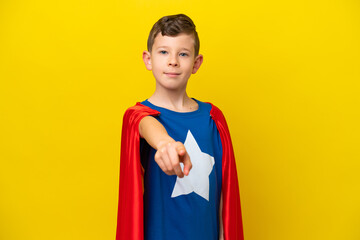 Little caucasian boy isolated on yellow background in superhero costume and pointing to the front