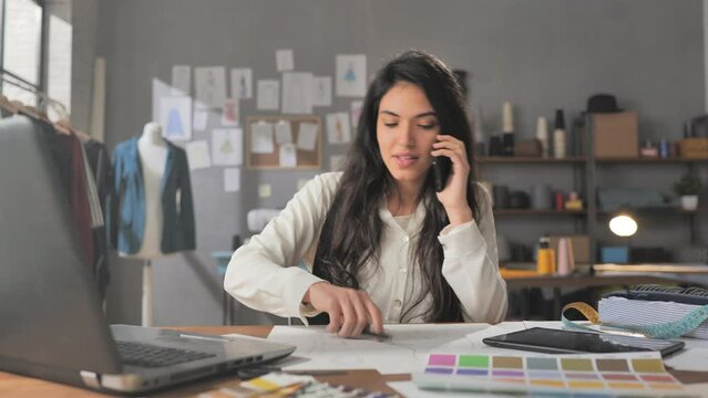 Young female fashion designer works in studio speaking at the phone takes online orders from customer using computer laptop while drawing clothes sketches in studio workshop