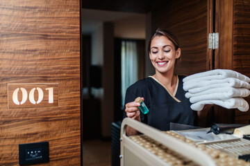 Beautiful young hotel chambermaid in uniform bringing clean towels and other supplies to hotel room. - 488181323