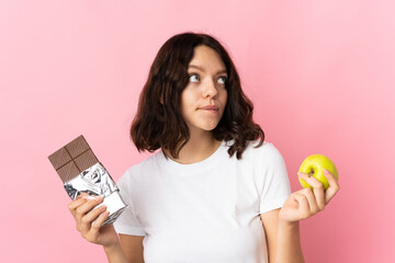 Teenager Ukrainian girl isolated on pink background having doubts while taking a chocolate tablet...