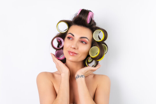 Beautiful housewife. Young cheerful woman with hair curlers and bare shoulders getting ready for a date night. Makes a hairstyle at home on a white background.