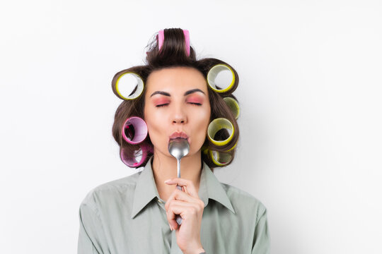 Beautiful housewife. Young cheerful woman with hair curlers, bright make-up, with a tablespoon in her hands, on a white background. Yummy, licks the spoon