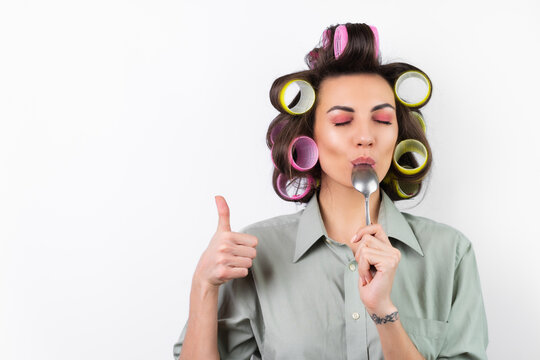 Beautiful housewife. Young cheerful woman with hair curlers, bright make-up, with a tablespoon in her hands, on a white background. Yummy, licks the spoon