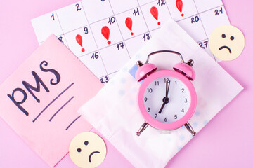 Calendar of women's menstruation, sad emoticons, Menstrual pads (sanitary napkin) in package, inscription of PMS on a piece of paper, an alarm clock on pink background. 