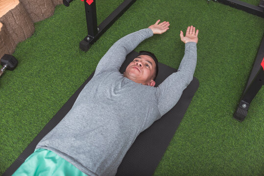 A fit asian man with arms extended, possible stretching or doing V-ups while lying on a black mat. Arms and legs raised. Working out his core at his home gym.